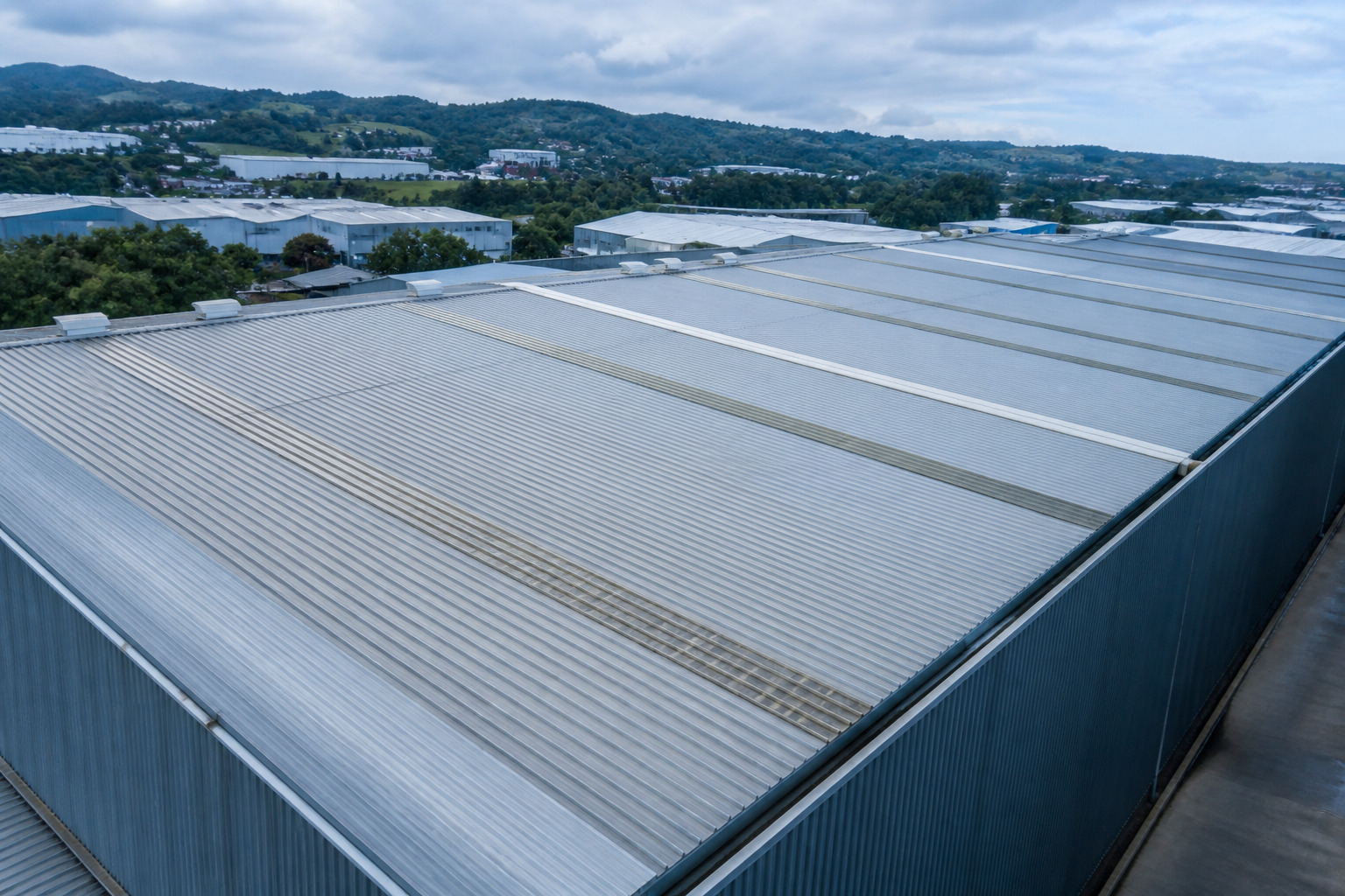 Elevated view across a large warehouse roof of profiled metal sheeting, with an industrial estate and hills in the distance